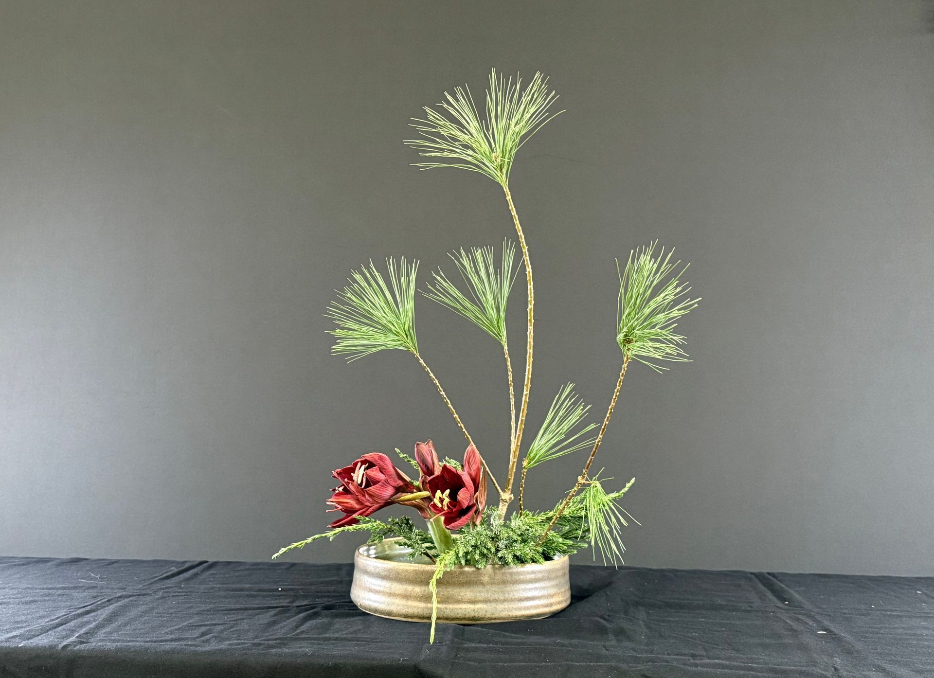 Bloemstuk: Rode bloemen, groene naalden en dunne stengels in een ronde vaas op een donkere ondergrond.