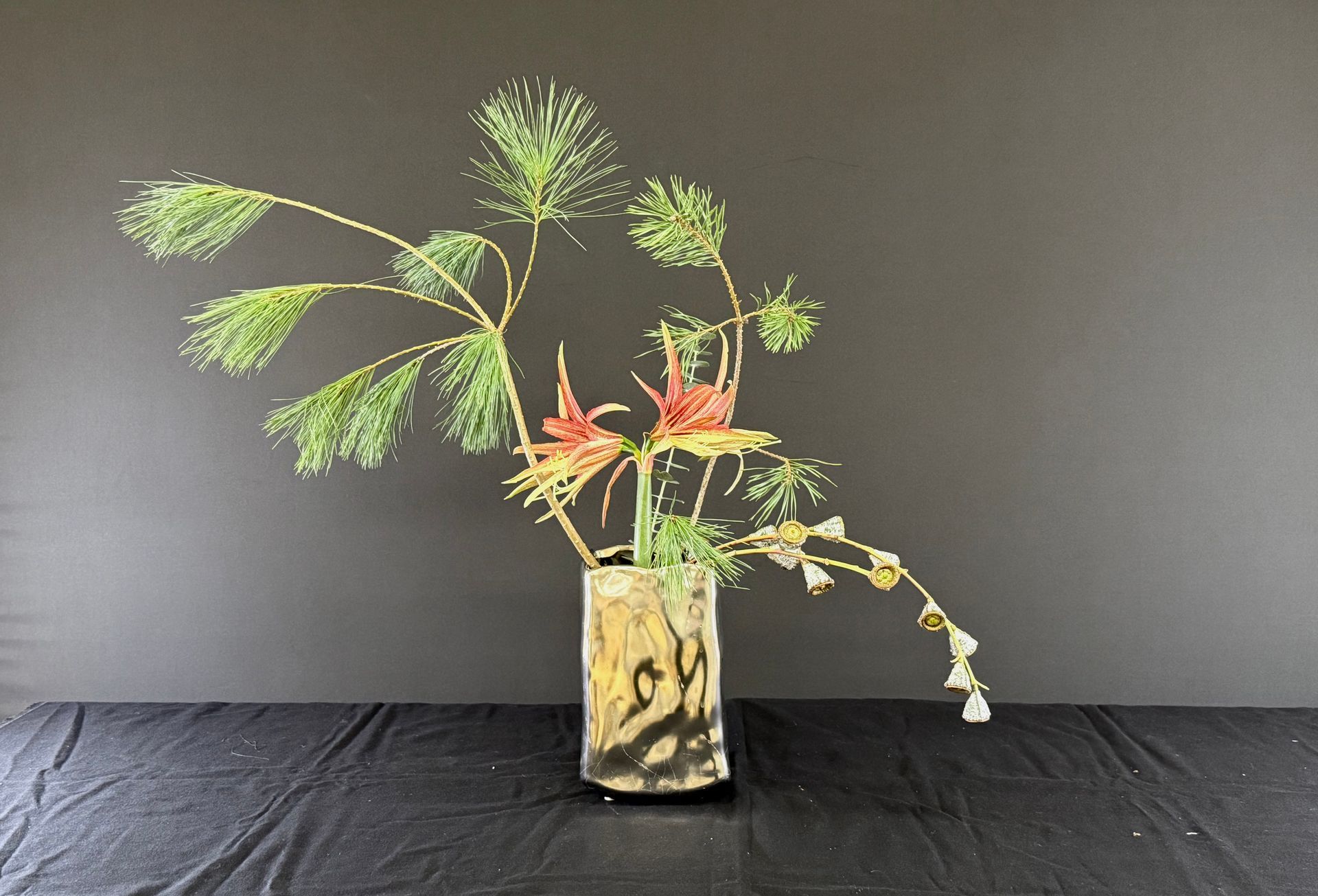 Bloemstuk in een cilindervormige vaas. Groene, rode en gele stengels en bladeren tegen een donkere achtergrond.