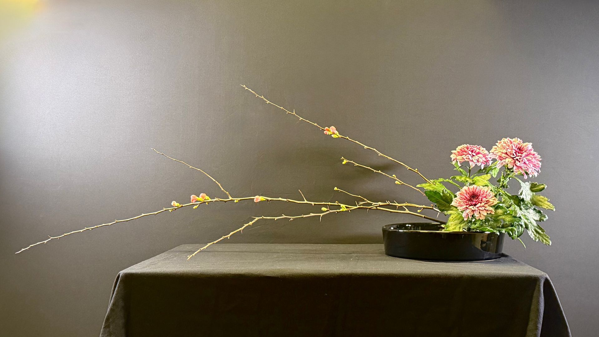 Bloemstuk op tafel: roze bloemen, slanke takken, donkere vaas, tegen een grijze achtergrond.
