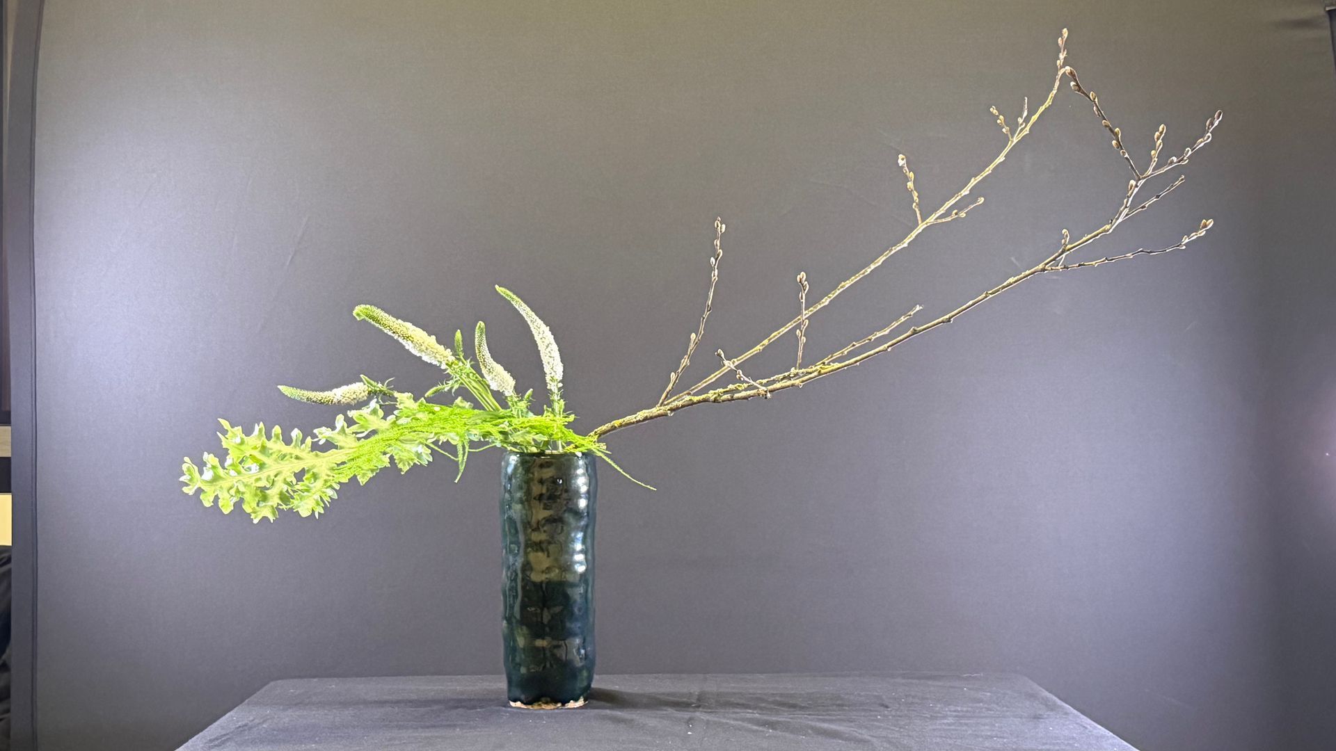 Bloemstuk in een donkere vaas tegen een grijze achtergrond. Met groene takjes en een tak.