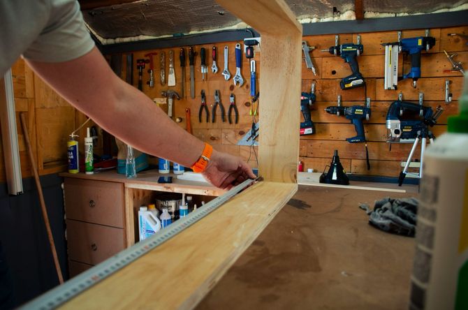 Person measuring a wooden board in a workshop with tools on the wall