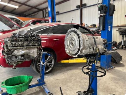 A car engine and transmission sit on separate mechanic stands inside a repair shop, with a red sedan parked behind them. | Rapid Auto and Truck Repair