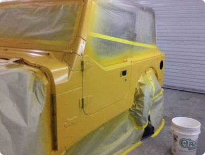 A yellow Jeep, partially masked with plastic and tape for painting, sits in a garage next to a bucket. | Rapid Auto and Truck Repair