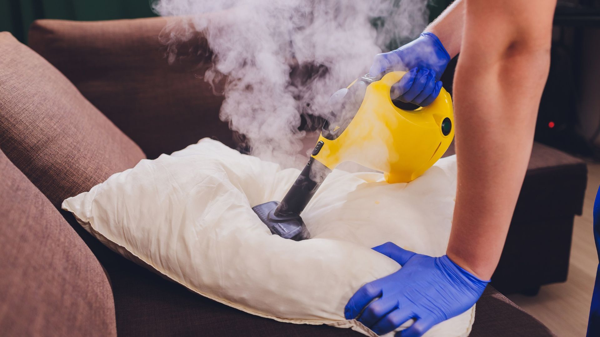 Person wearing blue gloves steam cleans a pillow with a yellow steam cleaner.