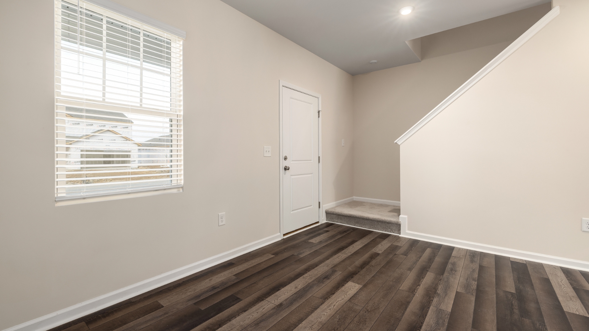 Empty room with dark wood floor, white door, window with blinds, and staircase.