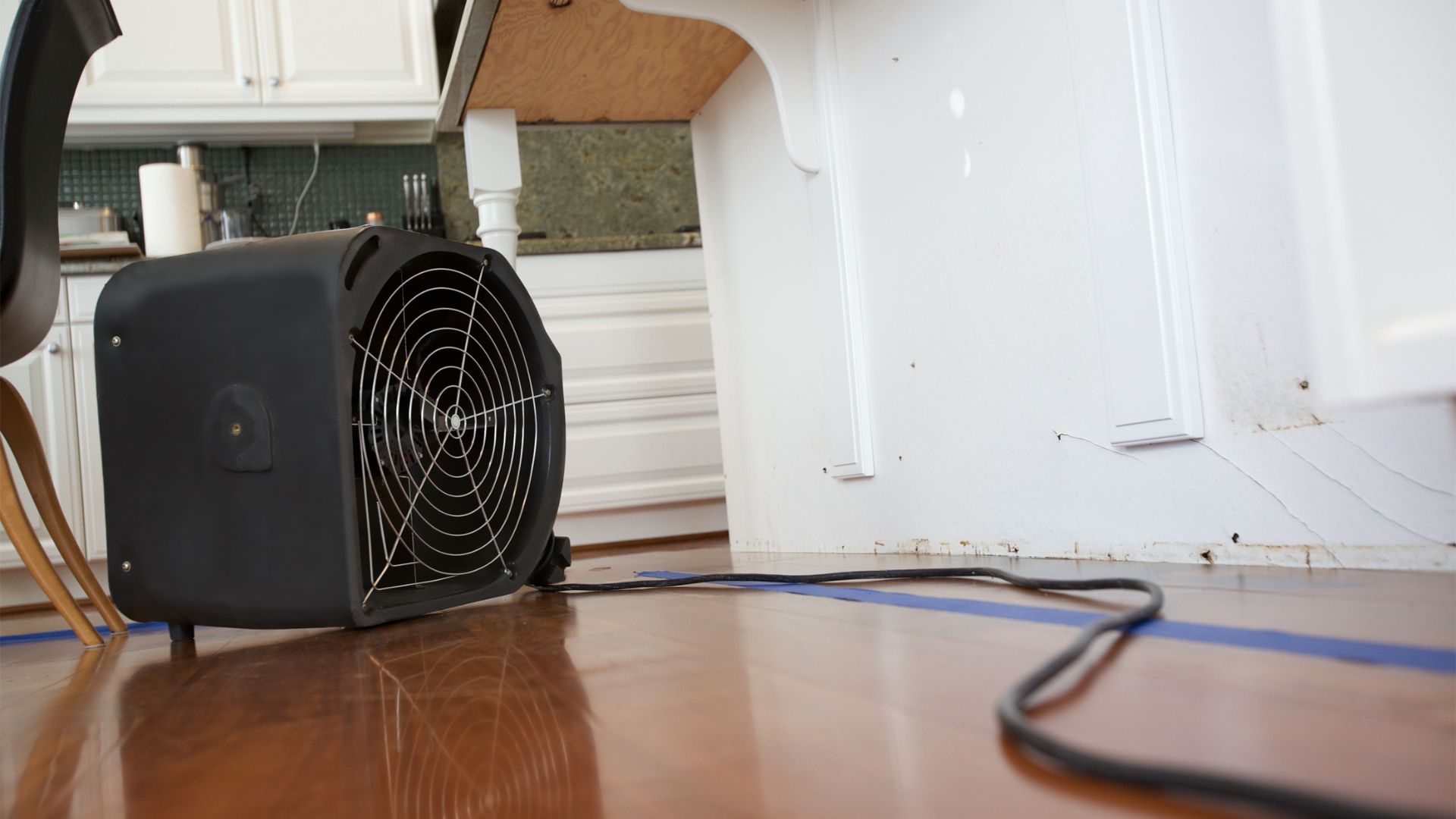 Black fan on wood floor near a white cabinet and chair, with a blue taped line.