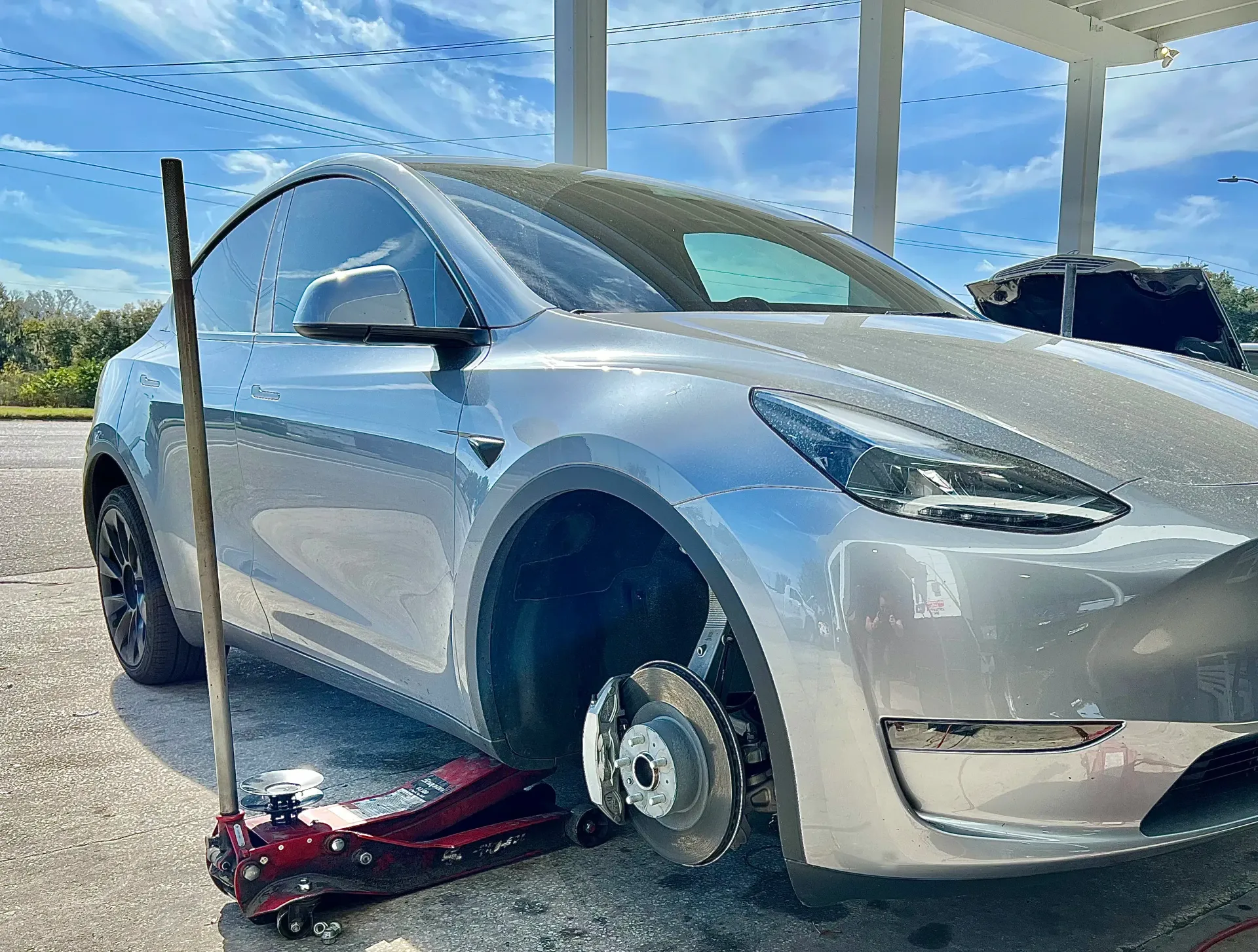A silver Tesla Model Y with its front passenger-side wheel removed, propped up by a red floor jack on a gravel surface | Infinity Tires and Auto Repair