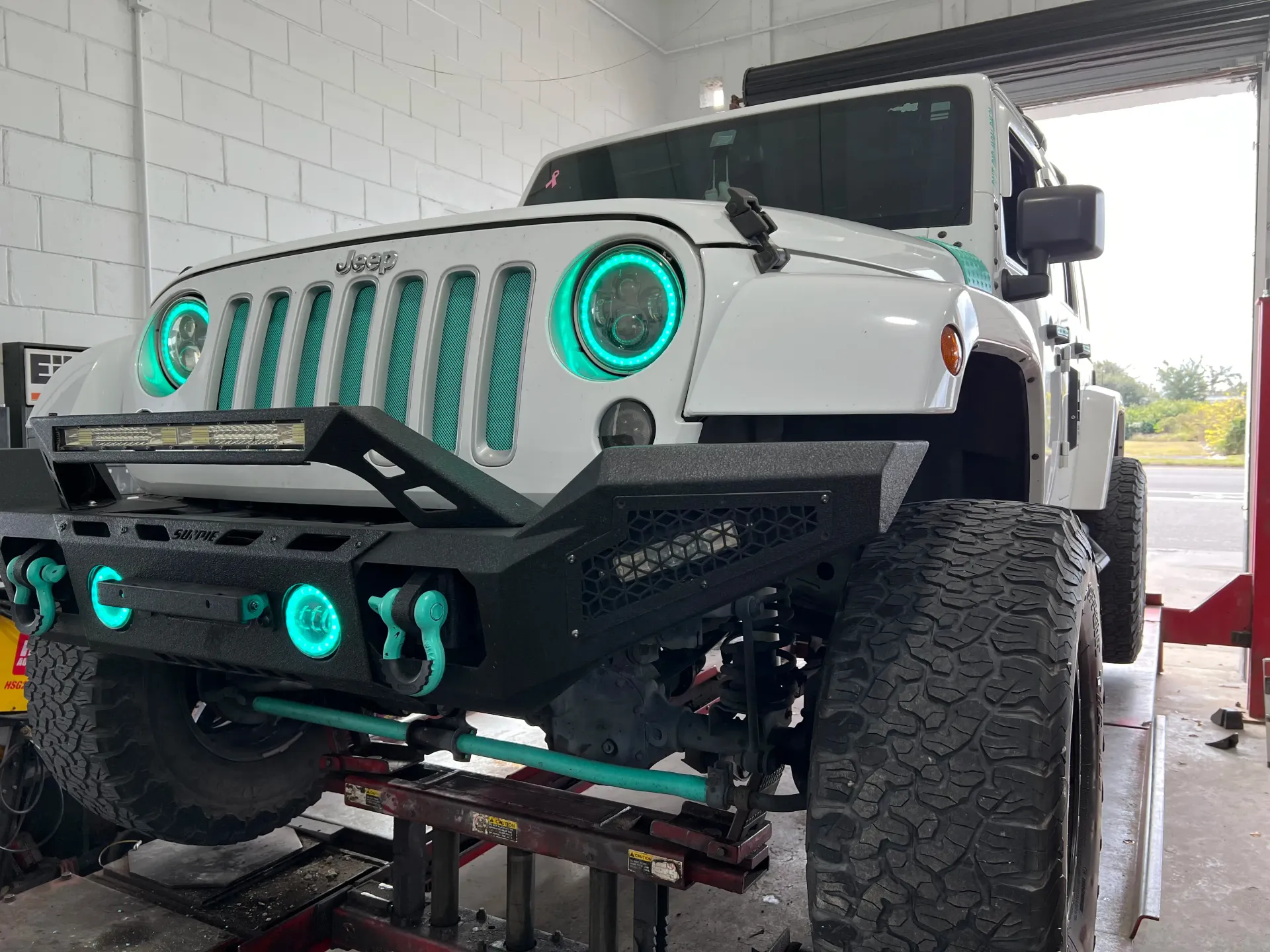 White lifted Jeep Wrangler with bright teal halo headlights and accents, parked on a hydraulic lift | Infinity Tires and Auto Repair