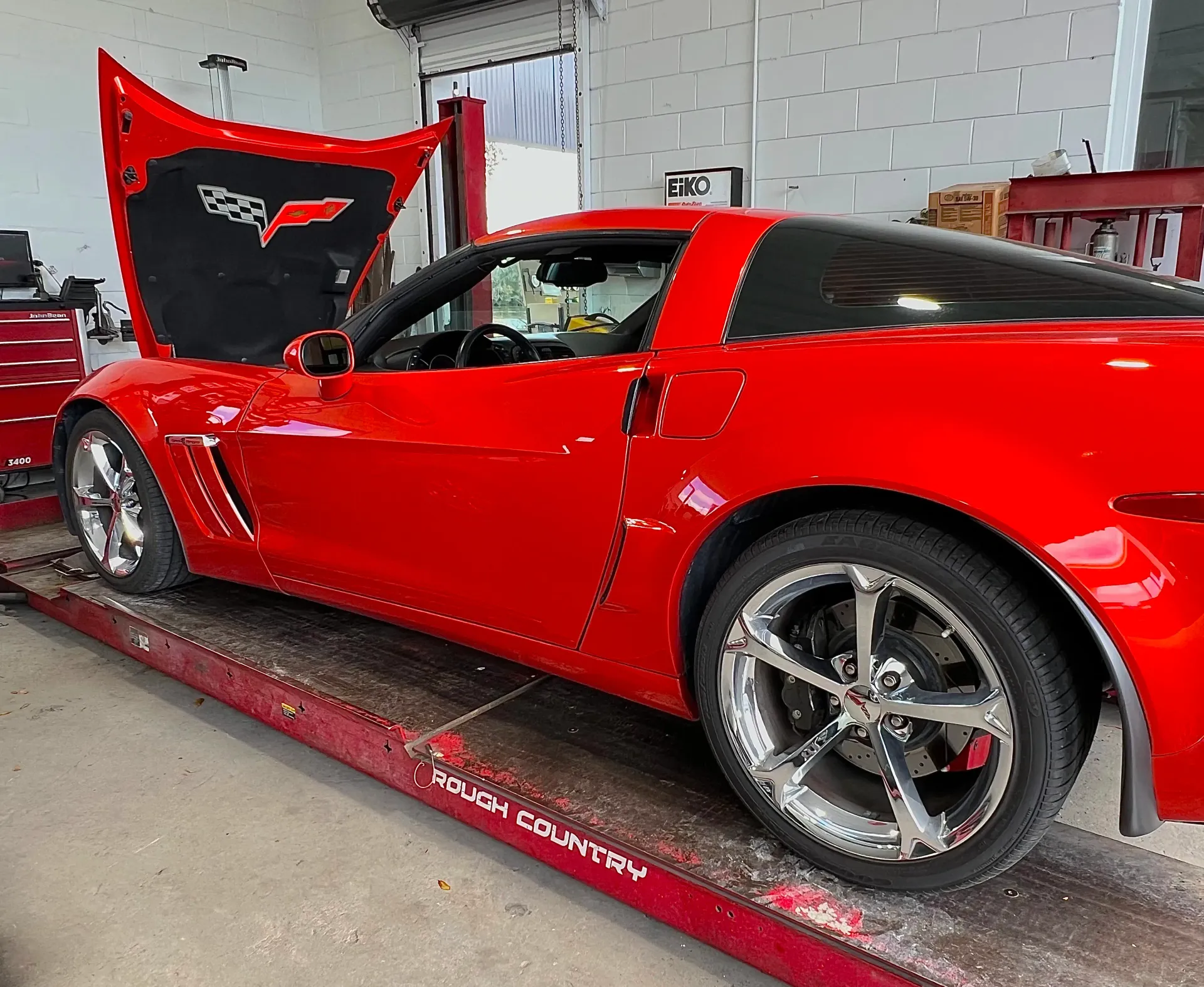 A bright red Chevrolet Corvette with its hood raised, parked on a mechanical lift | Infinity Tires and Auto Repair