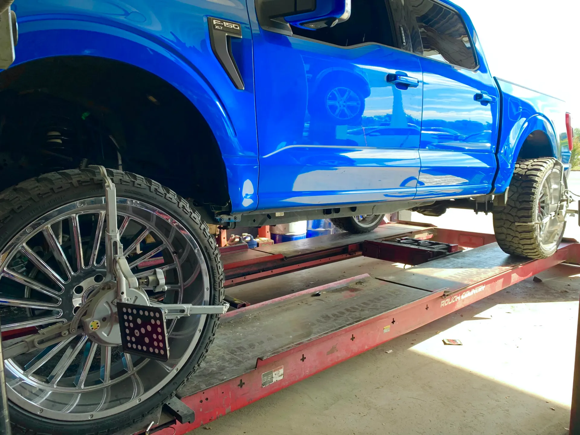 A blue truck on a lift for a wheel alignment, with a diagnostic sensor mounted to the front wheel | Infinity Tires and Auto Repair
