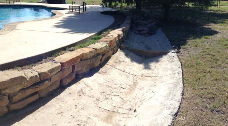 A stone wall surrounds a concrete walkway next to a pool.