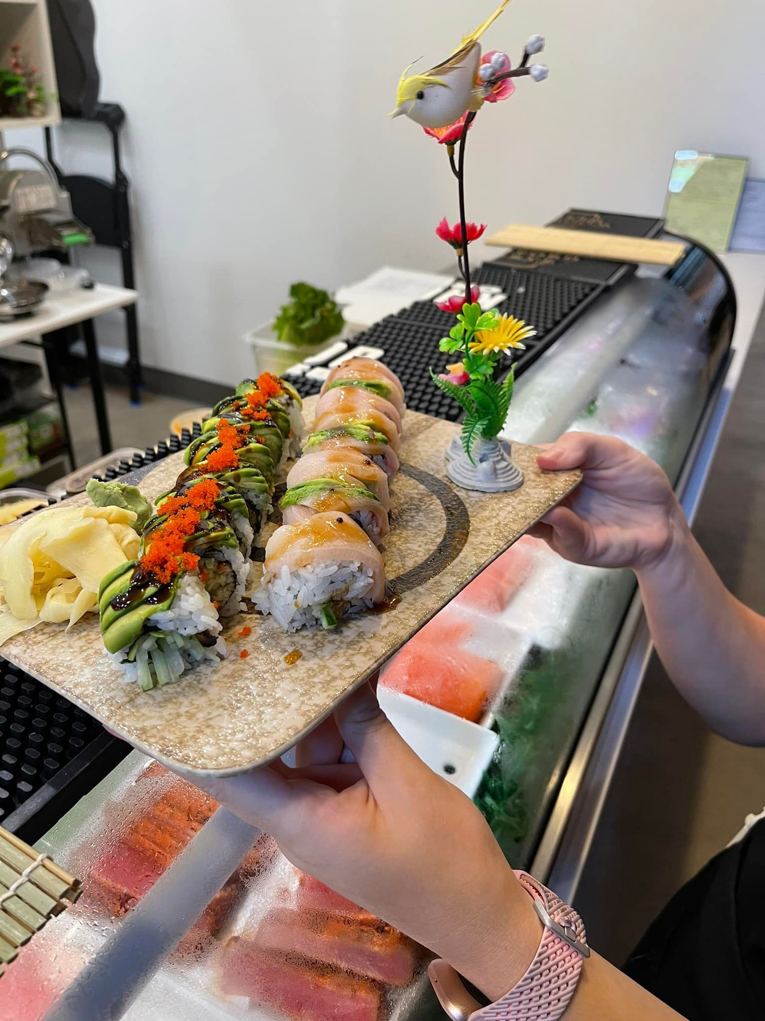 Sushi rolls on a decorative tray held by hands, near a sushi display. A small bird decoration sits on the tray.