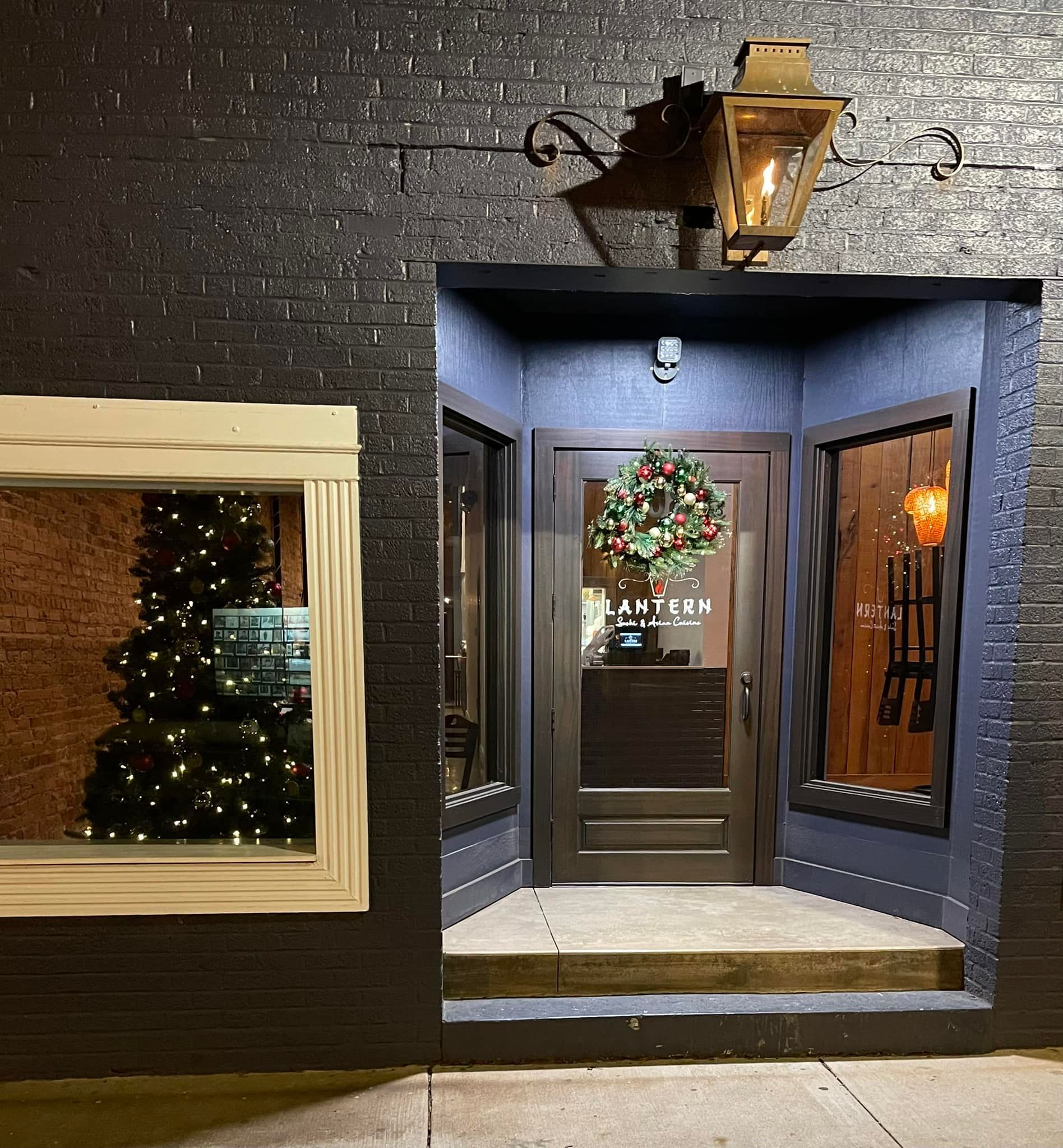 Black brick exterior with recessed doorway, wreath, and lit gas lamp. Window reflects Christmas tree.