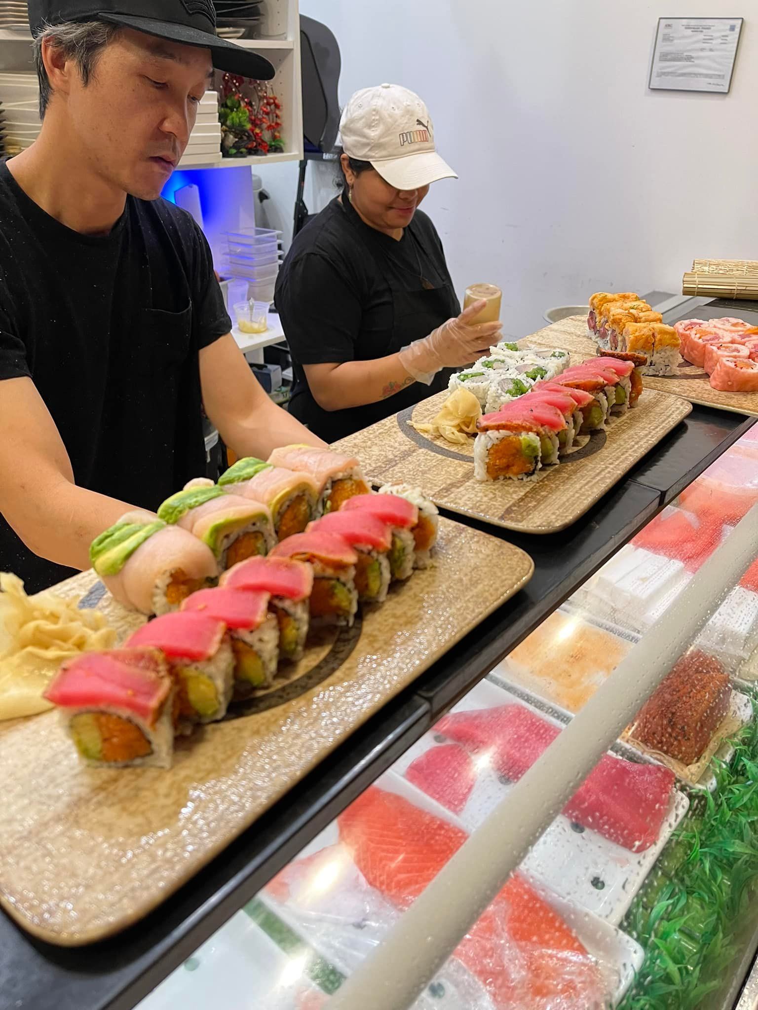 Two people preparing sushi behind a counter with rolls and ingredients on display.