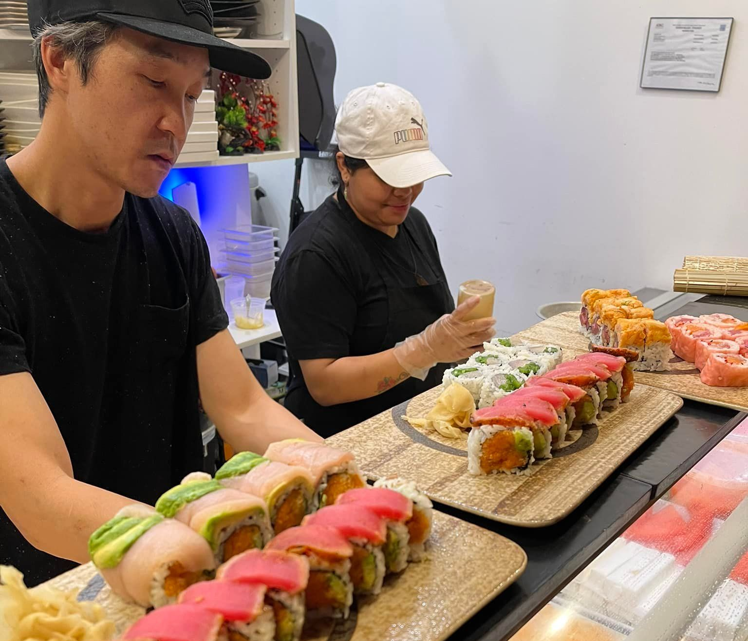 Two sushi chefs prepare rolls behind a counter filled with sushi.