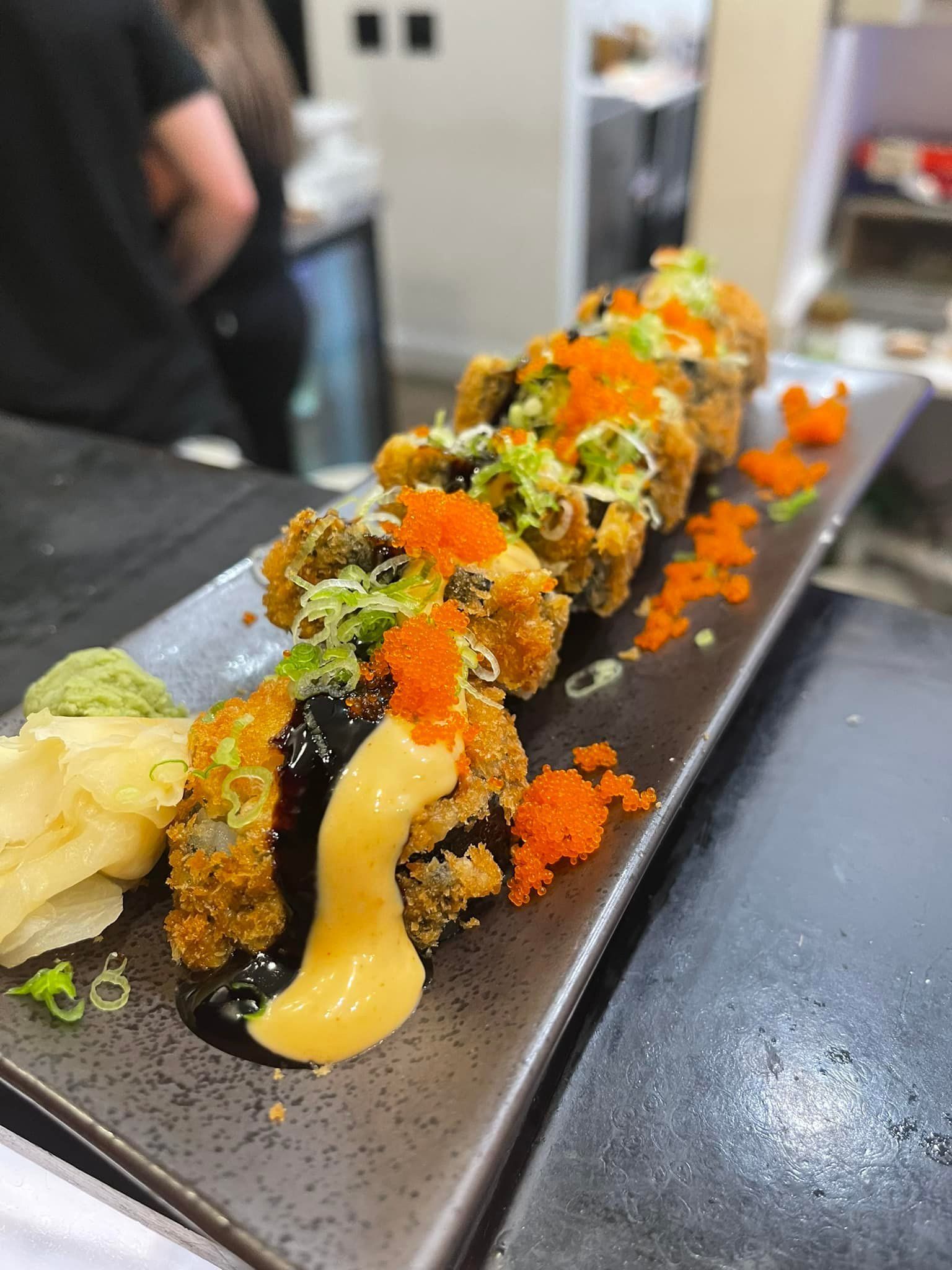 Fried sushi roll on a rectangular plate, topped with orange roe, sauce, and green onions.