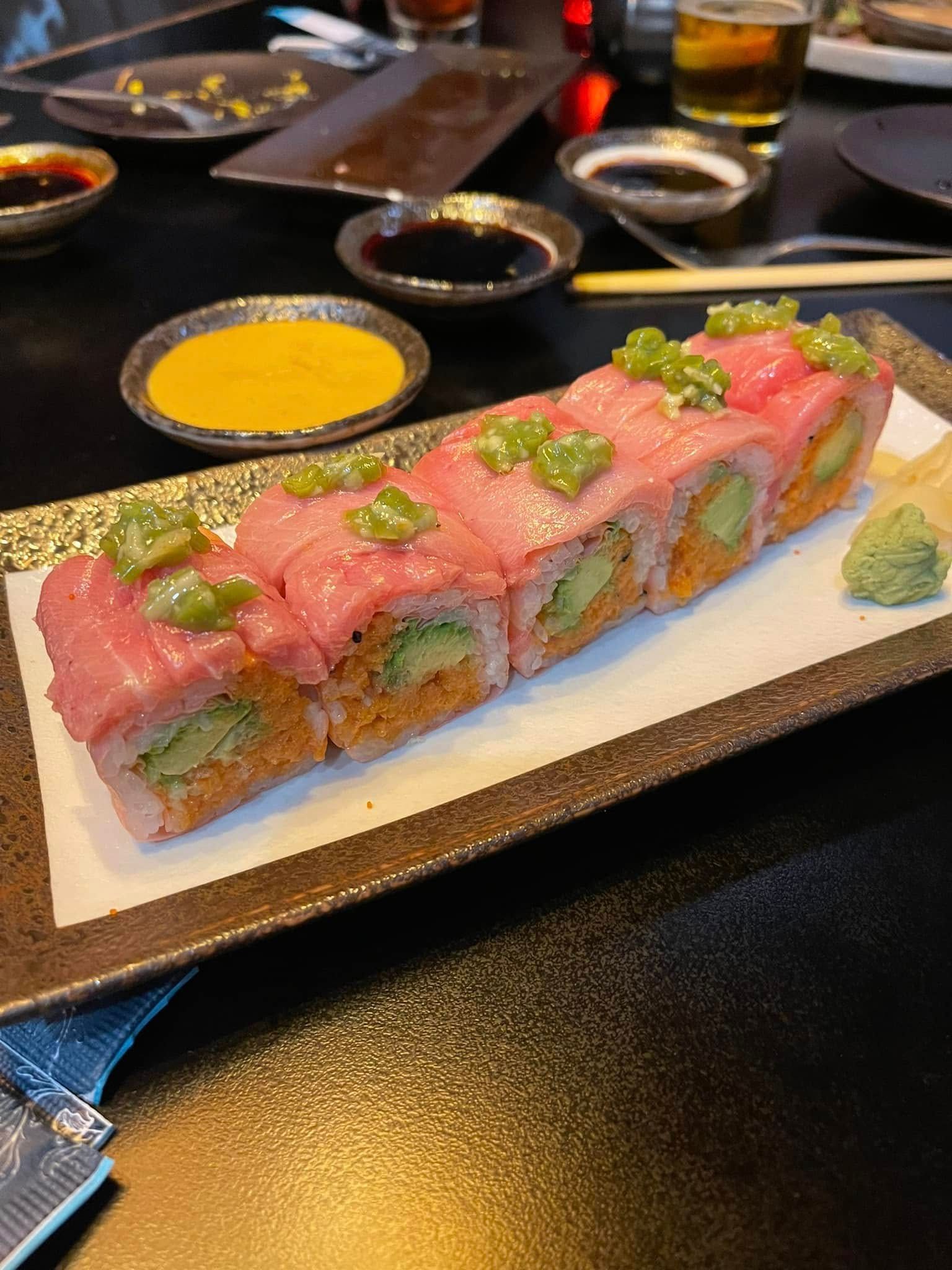 Sushi roll with pink fish, avocado, and sauce on a rectangular plate with dipping sauces.