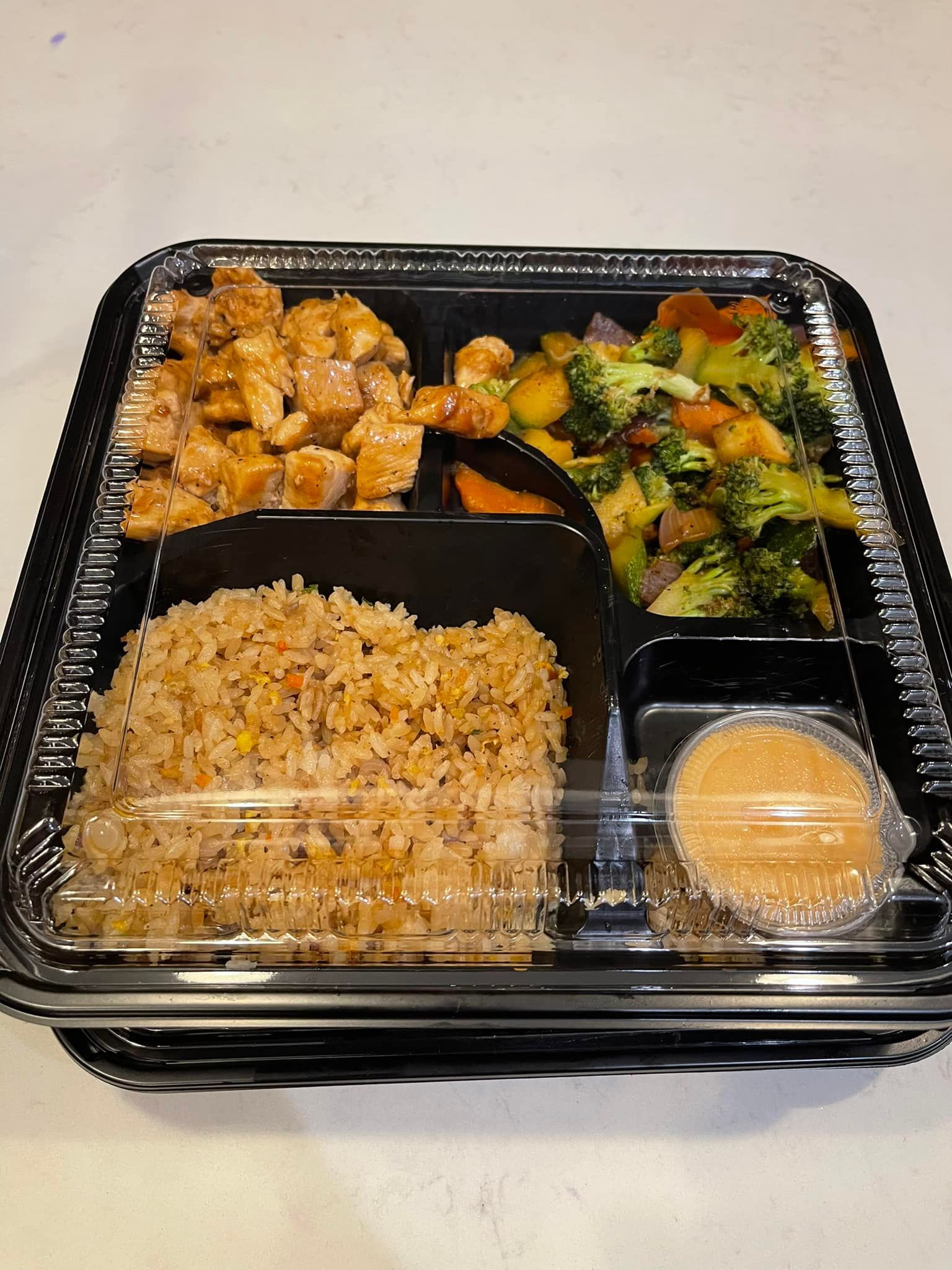 Bento box with chicken, fried rice, vegetables, and dipping sauce in a black container.