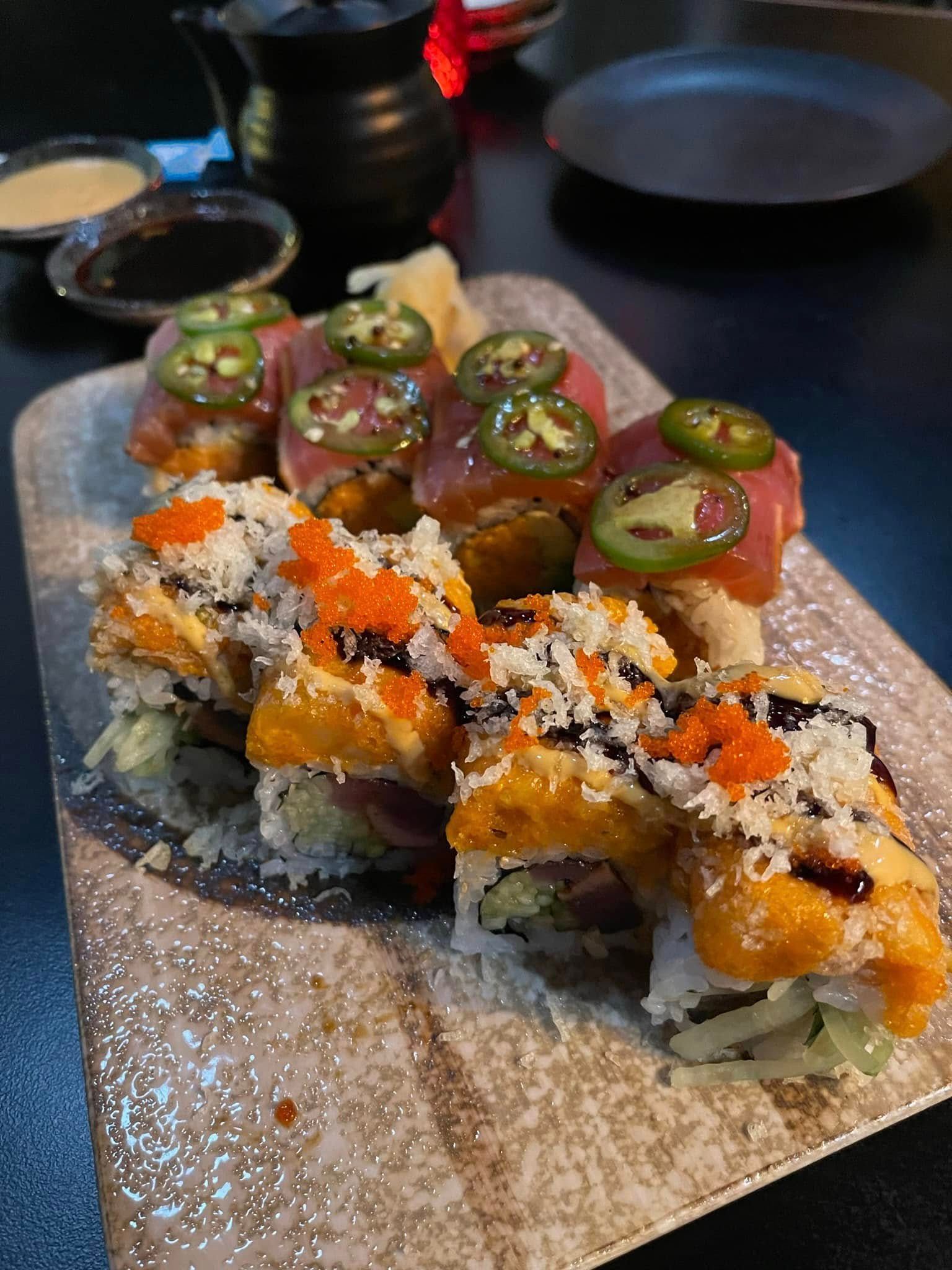 Sushi rolls topped with tuna, jalapeño slices, and orange roe on a textured rectangular plate.