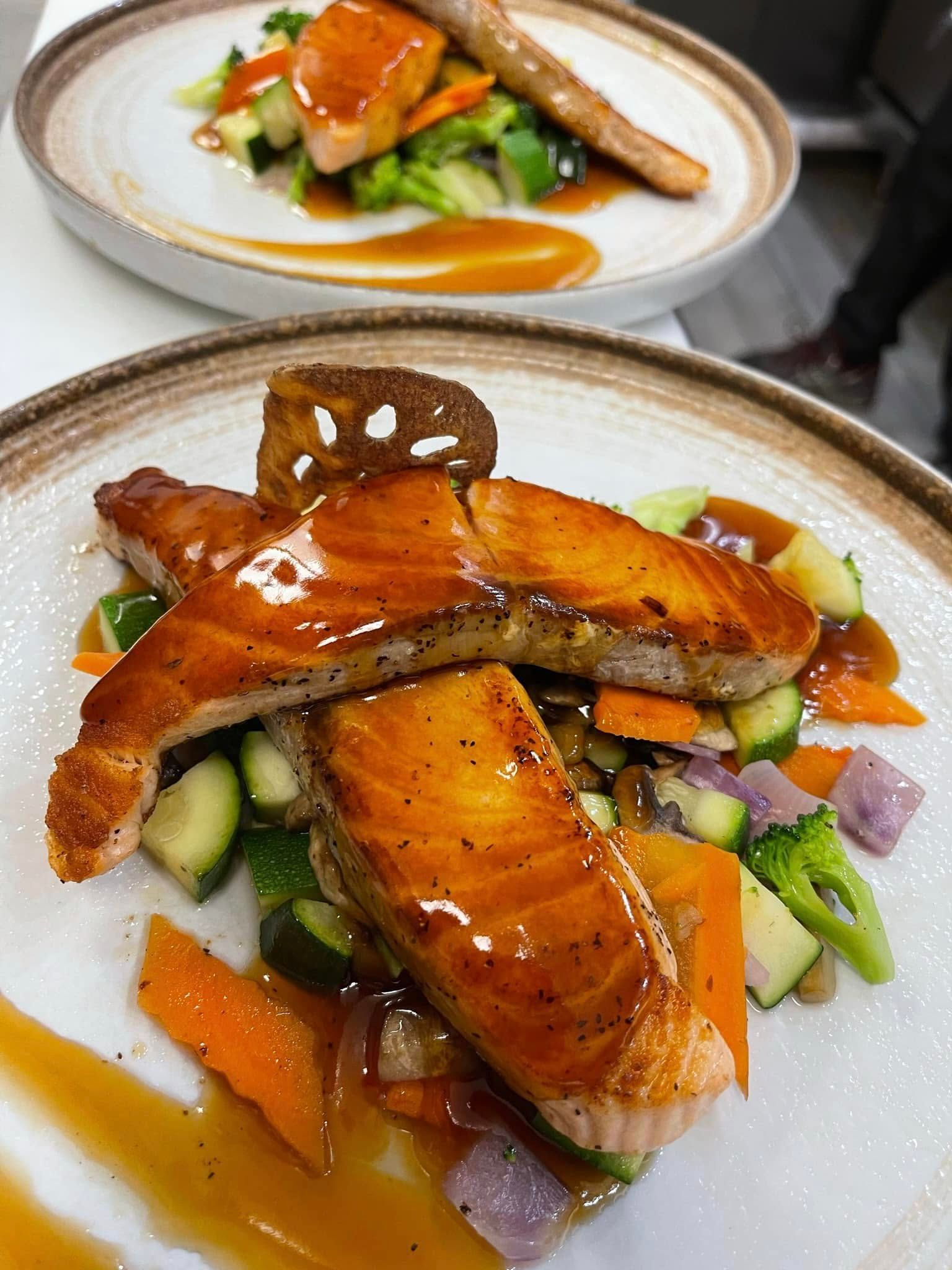 Two plates of glazed salmon with sauteed vegetables and sauce.