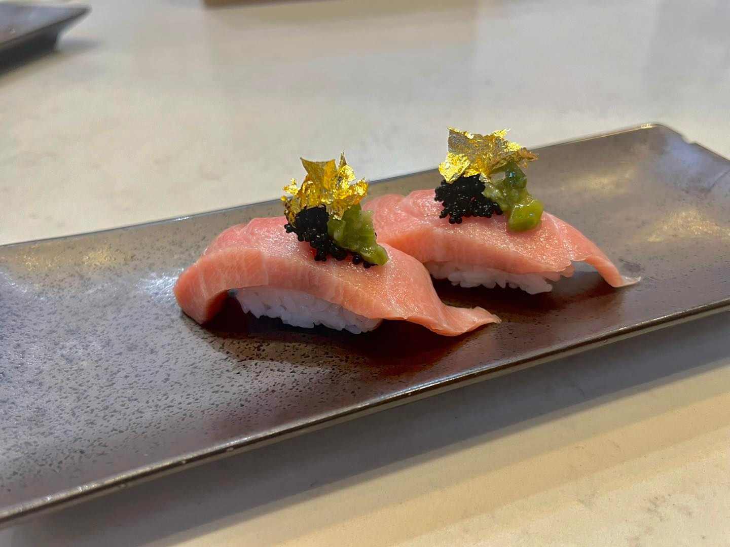 Two pieces of tuna nigiri sushi on a rectangular plate, topped with black caviar, green paste, and gold flakes.