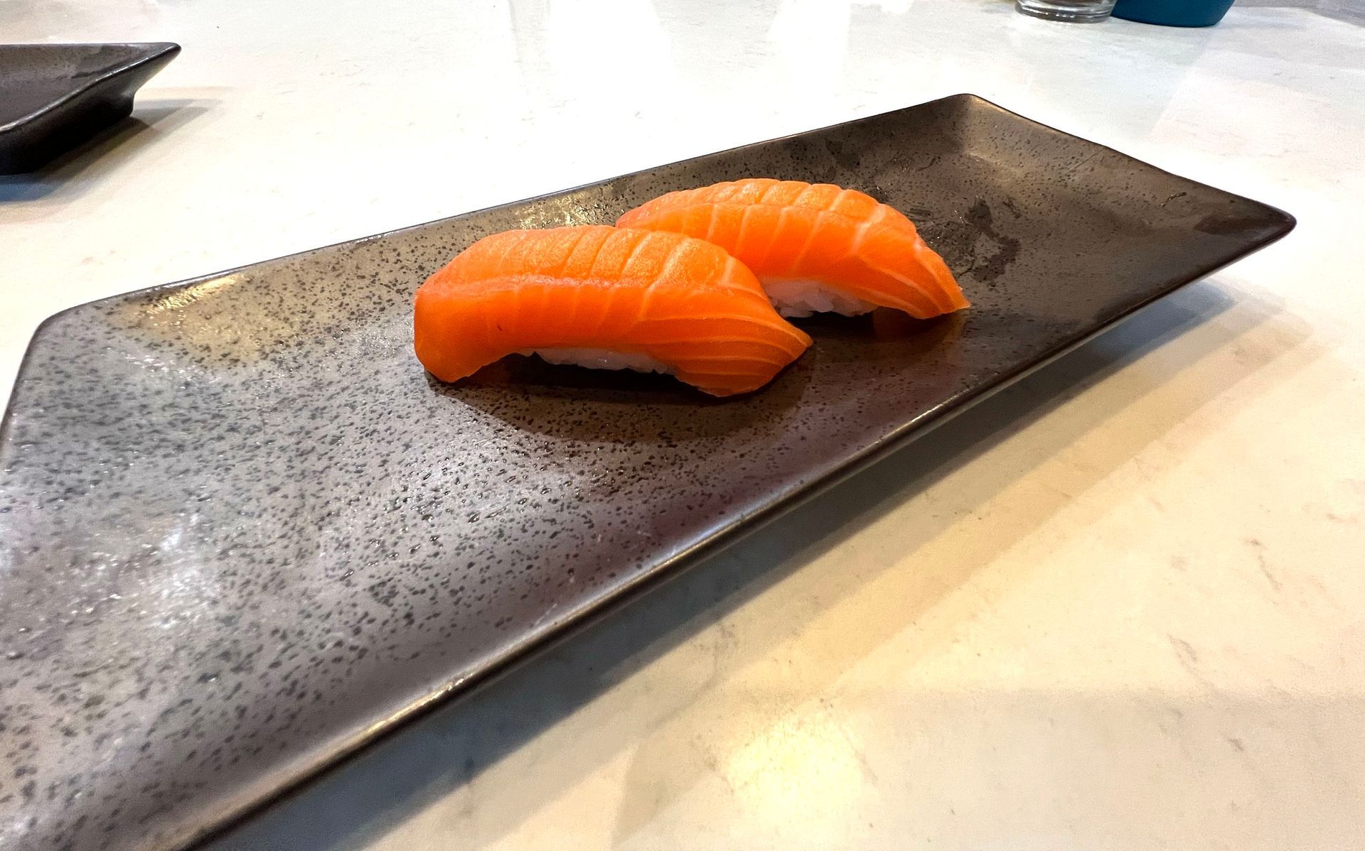 Two salmon nigiri sushi on a dark gray rectangular plate, set on a white table.