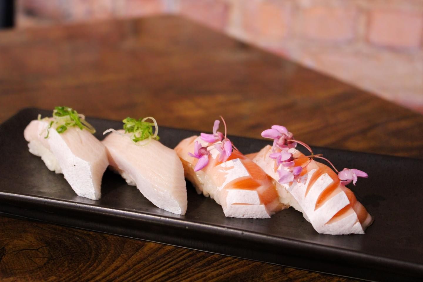 Sushi nigiri on a black plate, topped with green onions and pink flowers, on a wood table, against a brick wall.