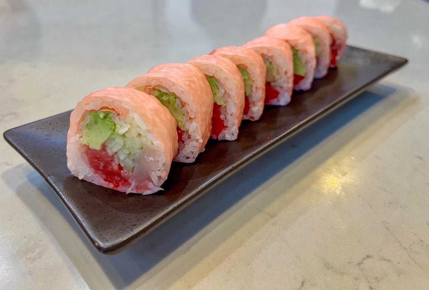 Sushi roll slices on a dark rectangular plate; pink, green, and red ingredients.