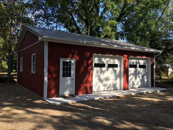 A red garage with two white garage doors is surrounded by trees.