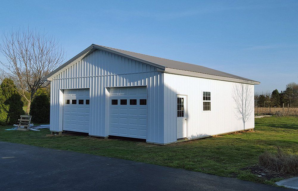 A white garage with two garage doors is sitting in the middle of a grassy field.
