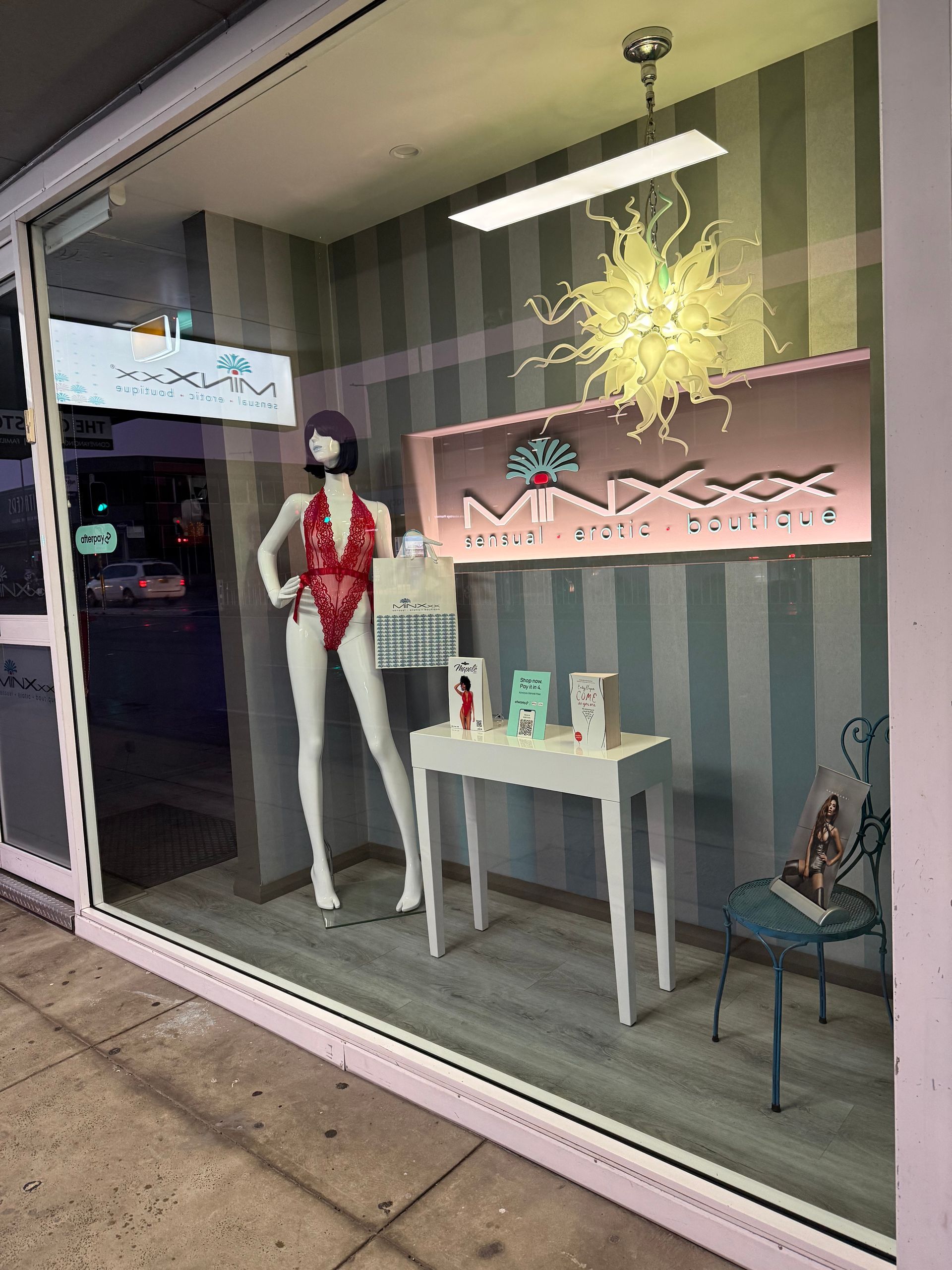 Window display: Mannequin in lingerie, boutique sign, white table with products. Striped wall.