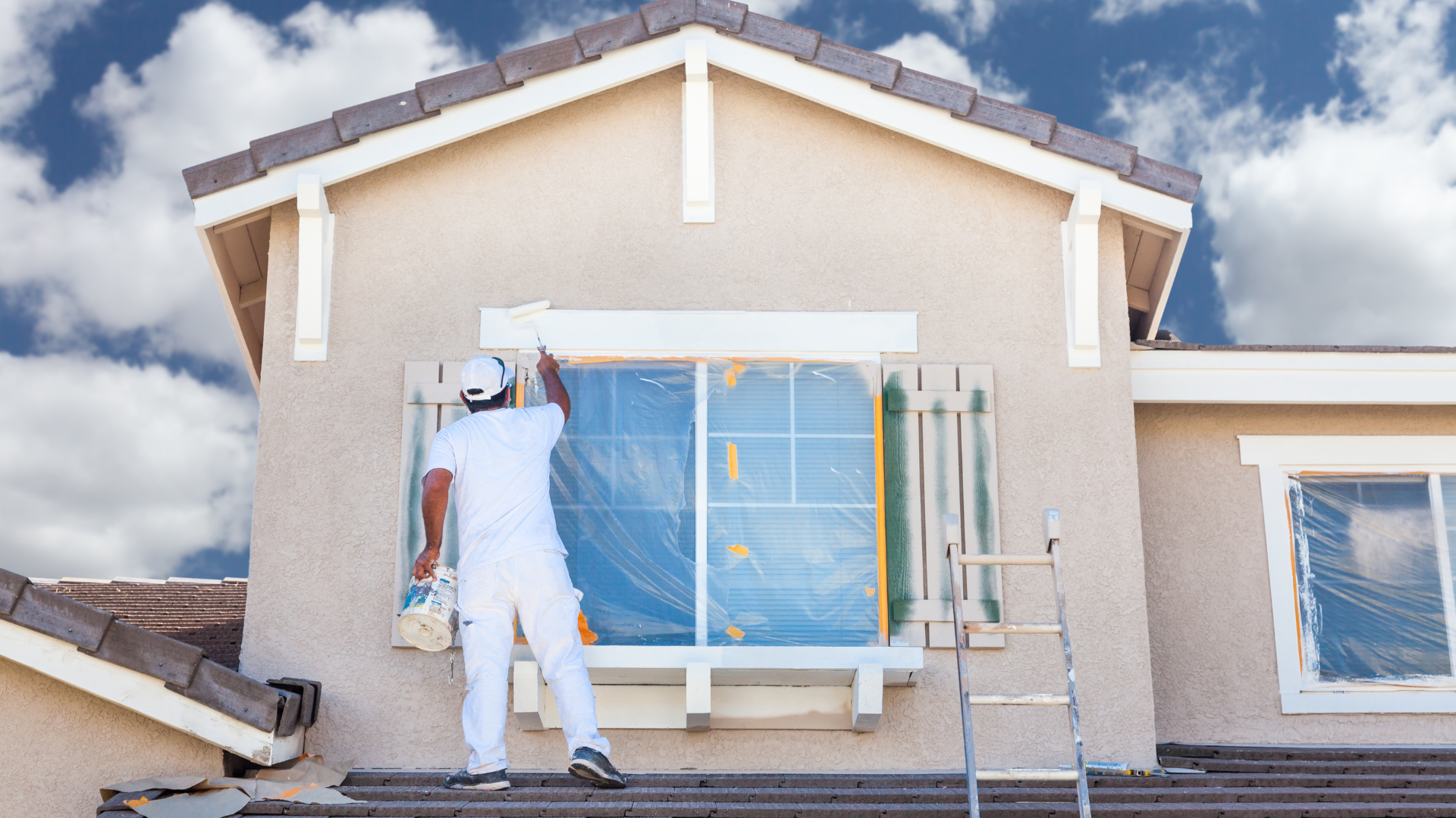 Painter in white overalls paints a house exterior, window covered with plastic, ladder nearby.