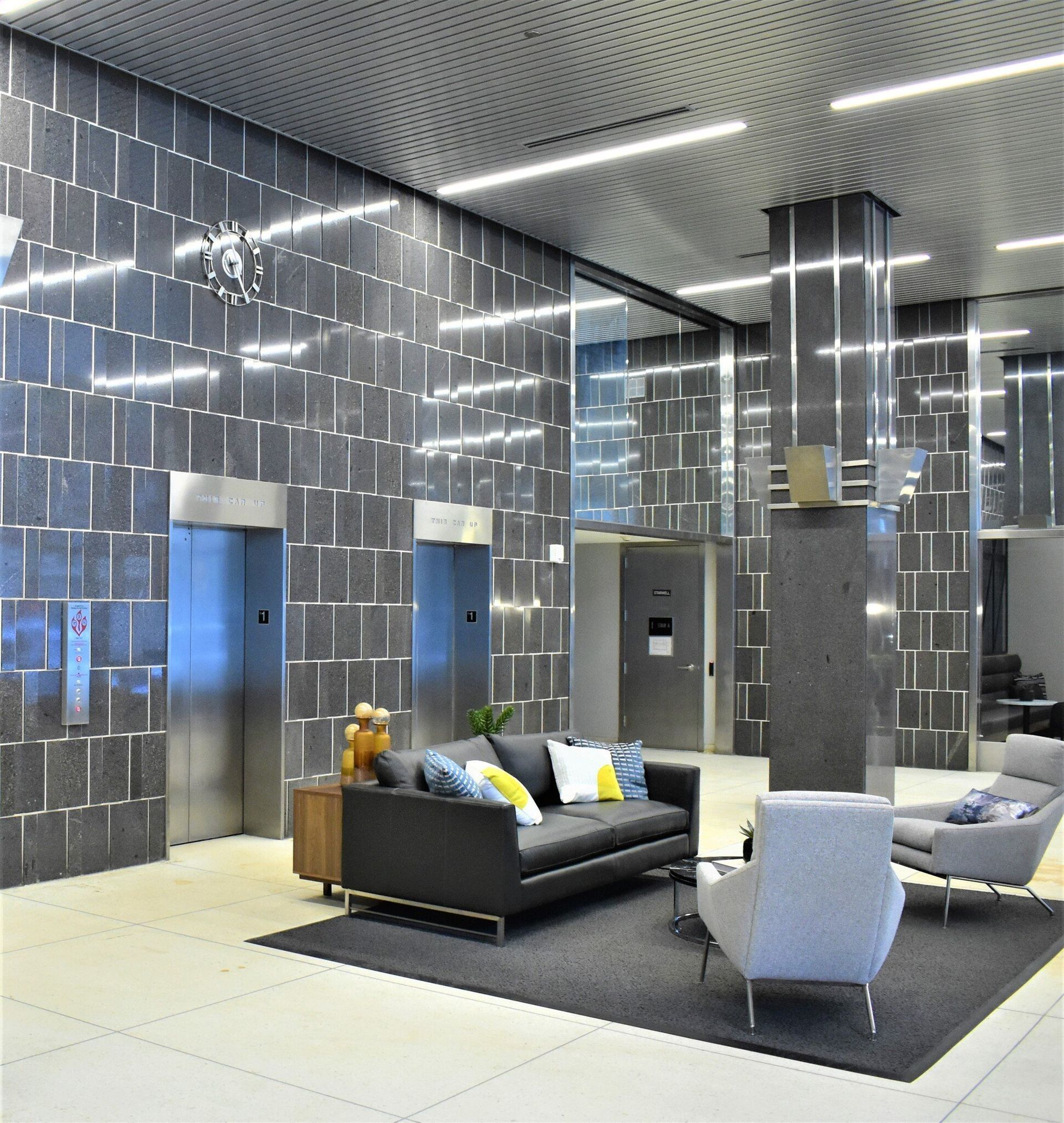 Modern apartment lobby with seating area and two elevator doors set in dark tiled walls.