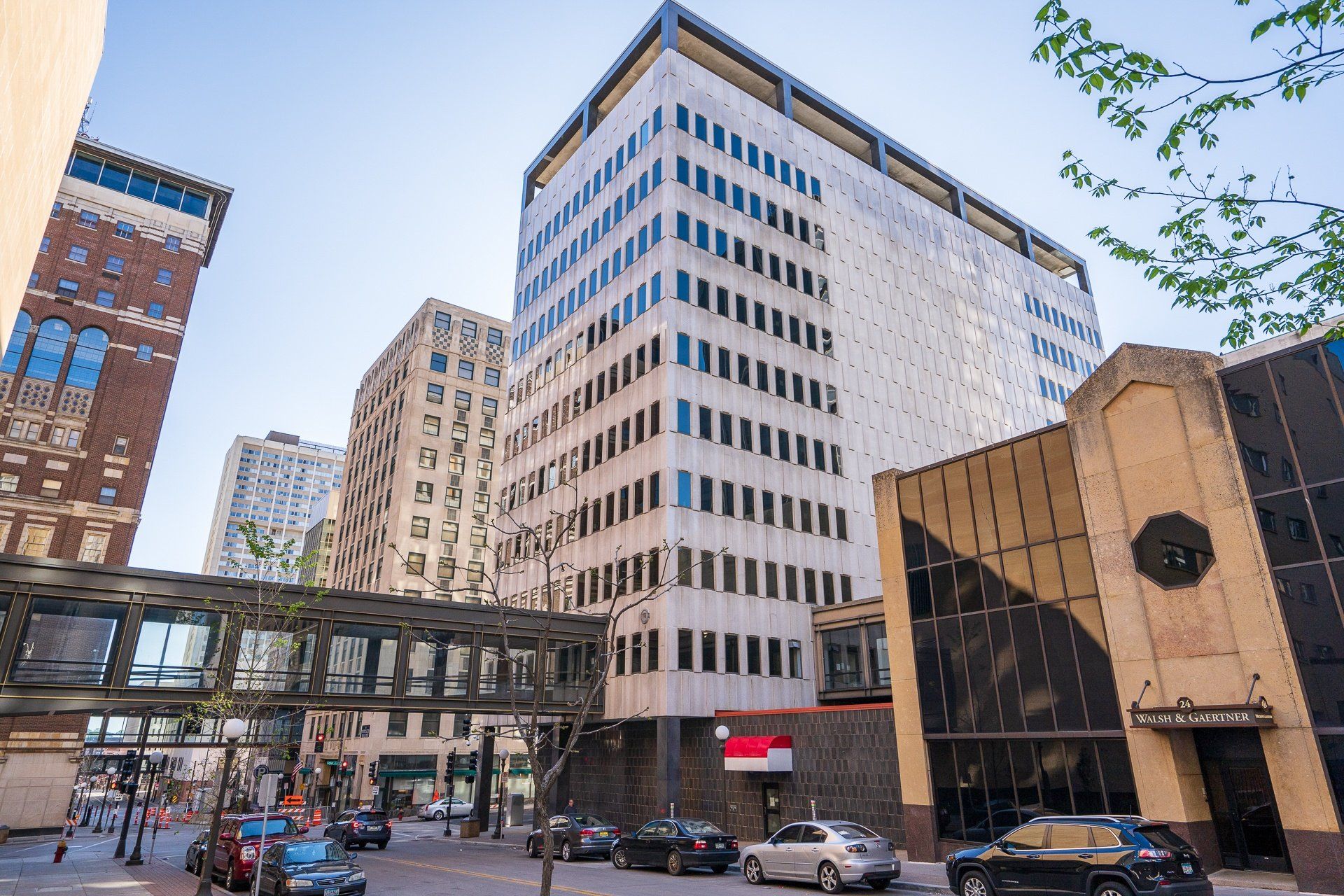 Urban street view of tall modern buildings with a glass-enclosed pedestrian skybridge.