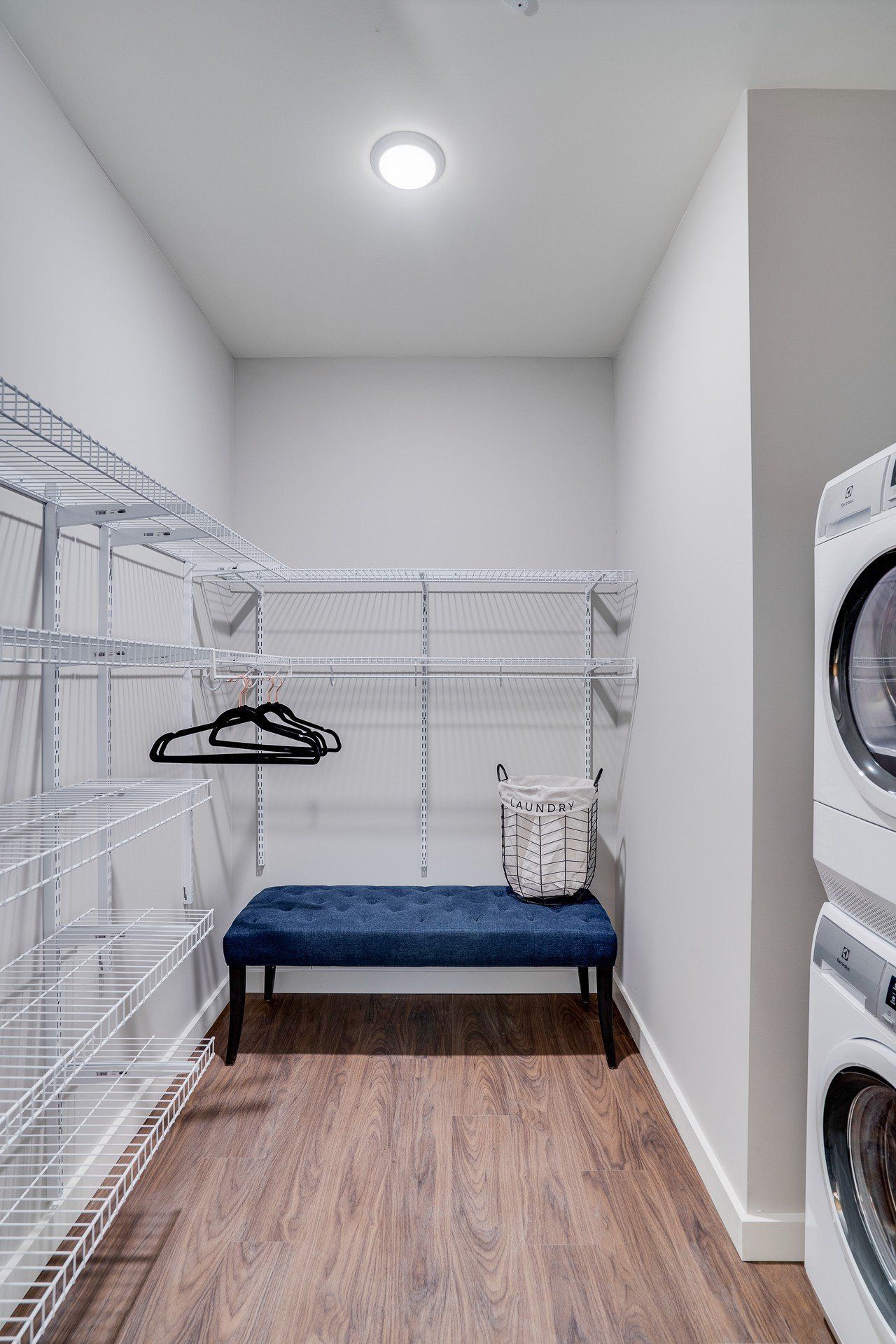 Laundry closet with white wire shelving, a blue bench, and a stacked washer/dryer on the right.