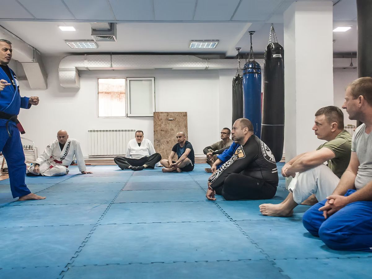 Jiu-Jitsu instructor in blue gi speaks to seated students at Ocean View Martial Arts in Norfolk, VA. Blue mats, various gis.