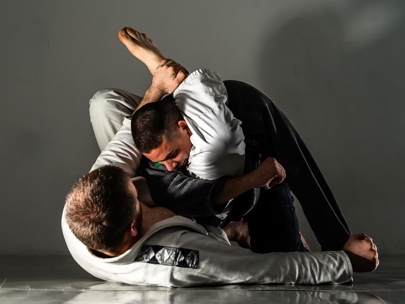 Two people in martial arts uniform grappling on a floor. One person is positioned on top in a dominant position.