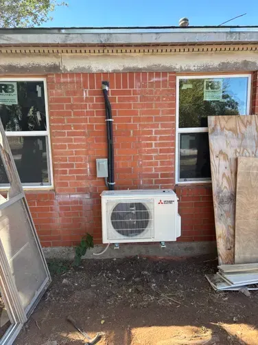 A white Mitsubishi mini-split outdoor HVAC unit mounted on a red brick exterior wall between two windows.