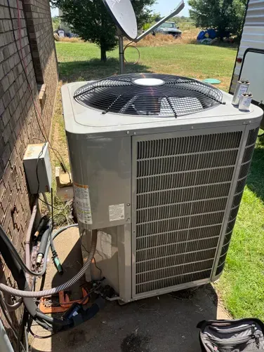 An outdoor central air conditioning unit sits on a concrete pad next to a brick house on a sunny day.
