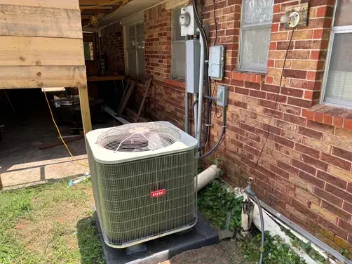 An outdoor HVAC unit sitting on a black pad next to a brick house wall with electrical conduit and a water spigot.
