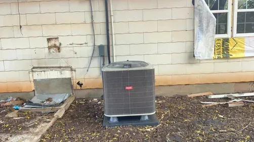 A grey Bryant outdoor air conditioning unit sits on a concrete pad against a block exterior wall near a construction site.