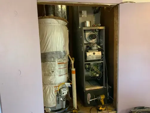 A water heater wrapped in a white blanket sits next to an open furnace unit inside a utility closet.