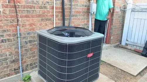 A Bryant central air conditioning unit sits on a concrete pad against a brick house wall near a person.