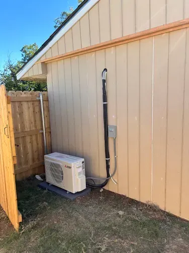 A beige Mitsubishi mini-split outdoor HVAC unit sits on a concrete pad against a tan exterior wall near a wooden fence.