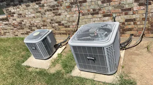Two gray outdoor air conditioning units sit on concrete pads against a brick exterior wall.