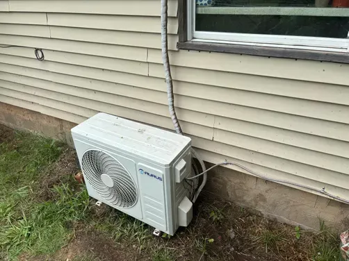 A white outdoor mini-split air conditioner unit sits on the ground against the beige siding of a house.