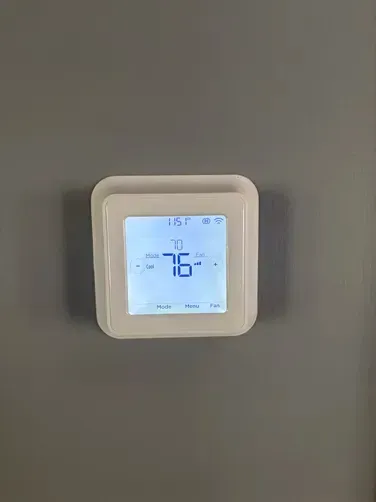 A white square thermostat mounted on a grey wall, displaying a room temperature of 76 degrees.