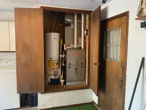 A utility closet with open wooden doors, containing a white water heater and a metal furnace inside a home setting.