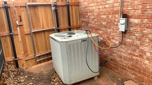 An outdoor air conditioning unit with gauges connected to its valves, sitting next to a brick wall and wooden fence.