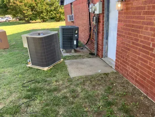 Two HVAC condenser units sit on the grass outside a red brick building with a utility door.